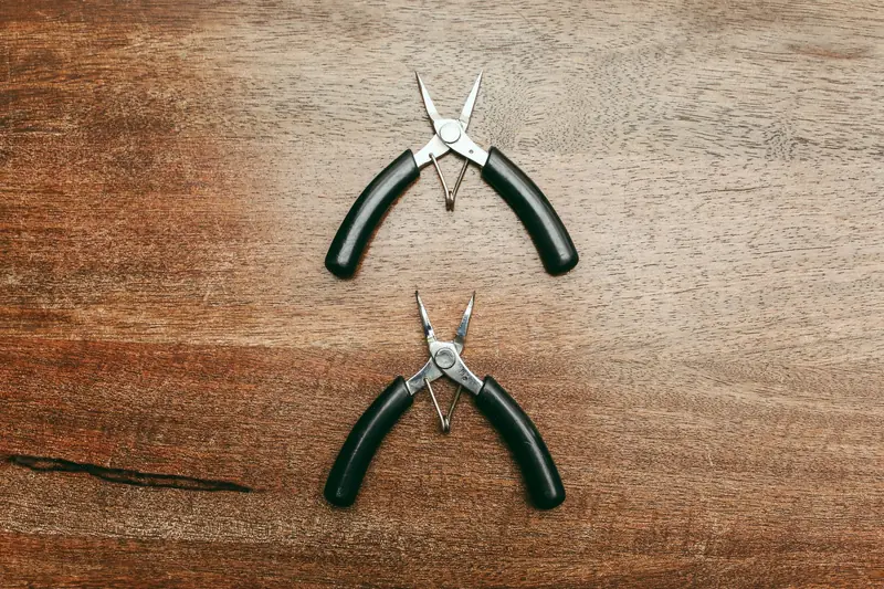Close-up of two needle-nose pliers on a wooden tabletop, showcasing stainless steel construction and black handles