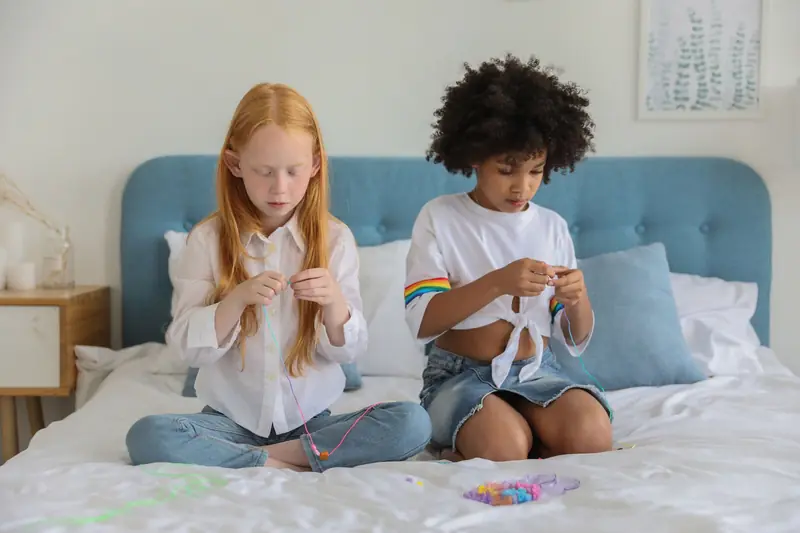 Concentrated multiethnic kids sitting on bed and making beads in cozy room in daytime