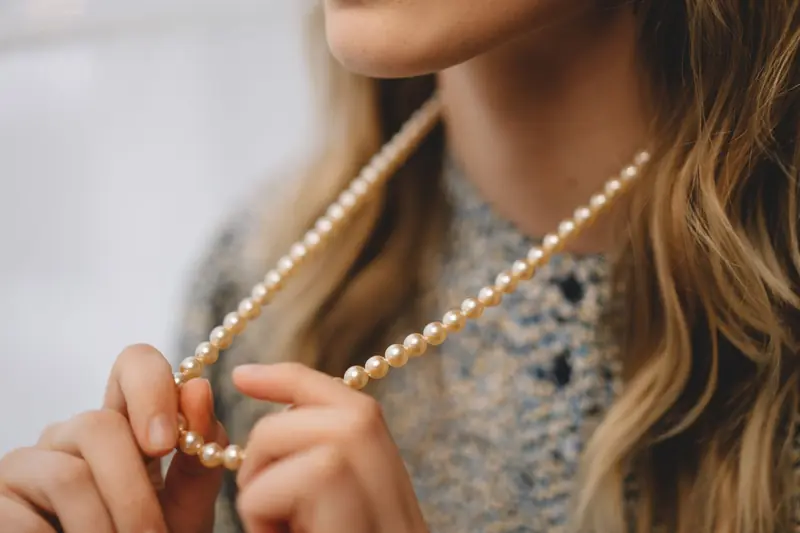A fashionable woman holding a pearl necklace with blonde hair visible in a stylish close-up shot