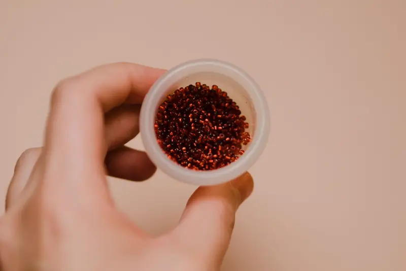 Close-up of a hand holding a round container filled with small red beads, perfect for crafts