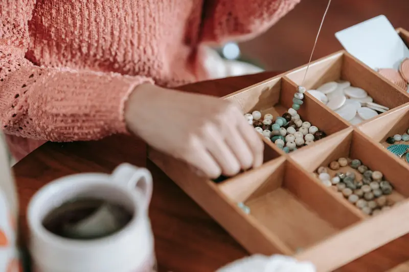 Crop unrecognizable female in casual outfit sitting at wooden table with cup of tea threading multicolored decorative beads and natural stones on fishing line w